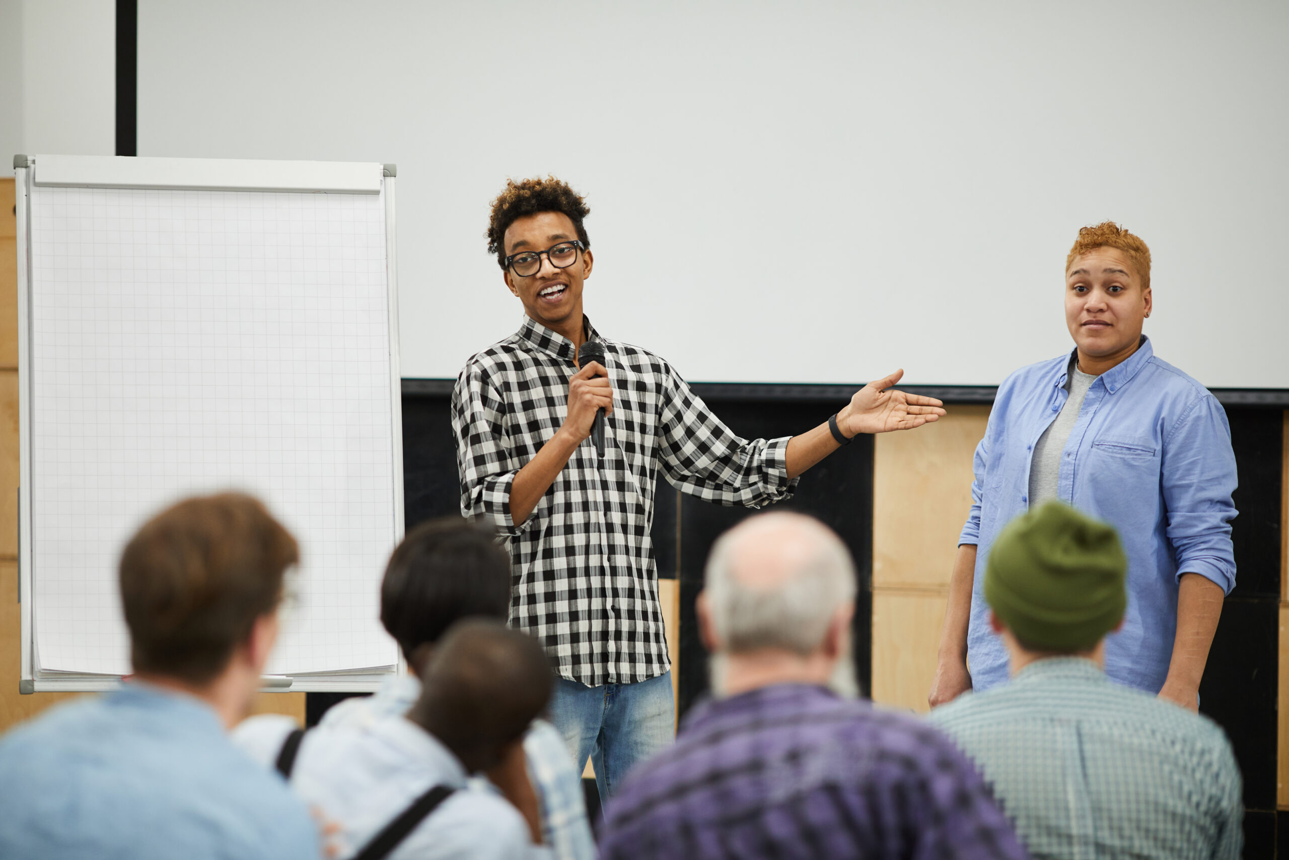 Two young men present at the front of a classroom; one holds a microphone and speaks while the other stands nearby, addressing seated attendees with a whiteboard beside them.